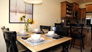 Un comedor con una mesa dispuesta para cuatro personas, rodeada de sillas de madera, un florero con girasoles y un cuadro en la pared.