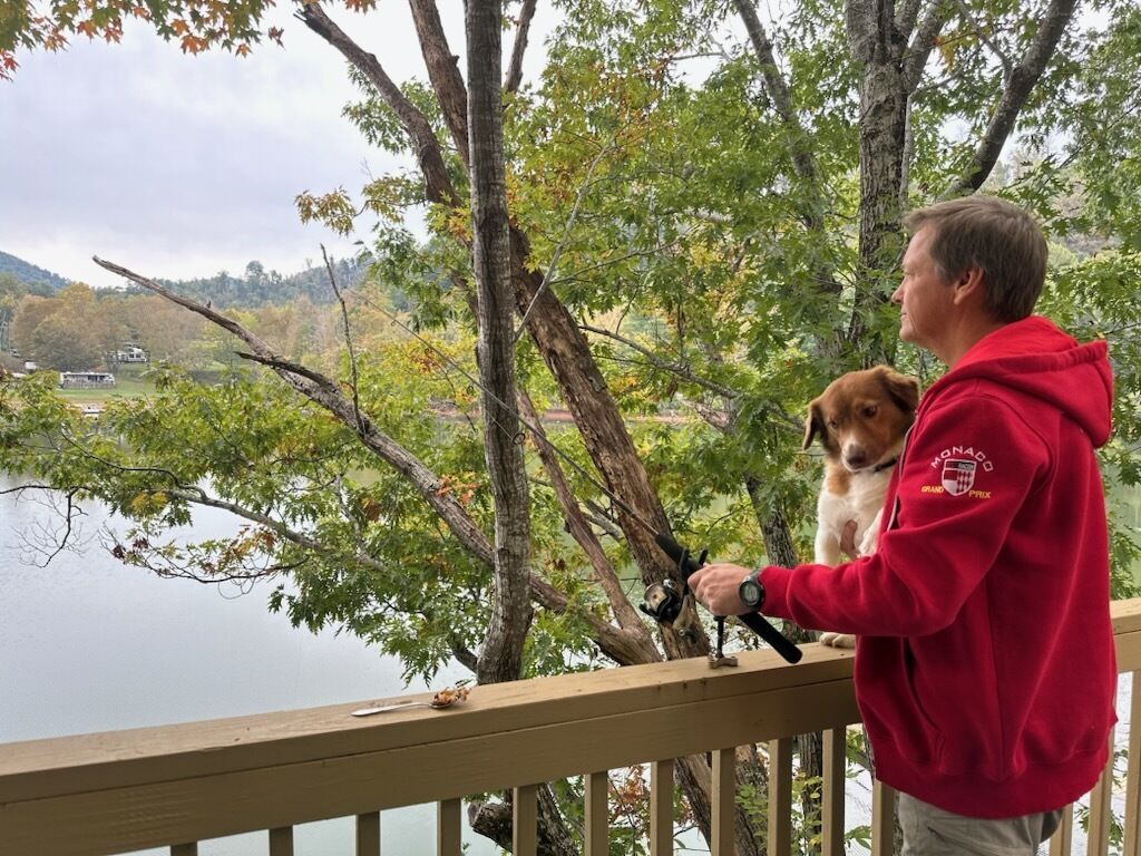 Una persona con sudadera roja parada en una terraza de madera con vista a un lago, árboles y colinas al fondo.