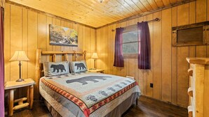 A wooden-paneled bedroom with a bed featuring a bear-themed bedspread, two bedside lamps, and a window with purple curtains. - Riverfront Cabin, Mountain Views, Fishing, and Rafting! (Sylva)