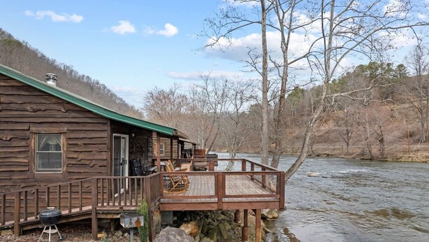 A rustic cabin with a deck overlooking a river, surrounded by trees. - Riverfront Cabin, Mountain Views, Fishing, and Rafting! (Sylva)