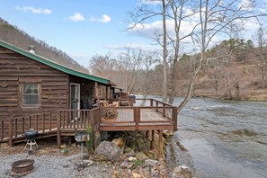A rustic cabin with a deck overlooking a river, surrounded by trees.