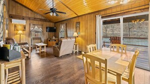A spacious room with wooden floors, a ceiling fan, a dining table with chairs, a leather sofa, and a large window overlooking a river. - Riverfront Cabin, Mountain Views, Fishing, and Rafting! (Sylva)