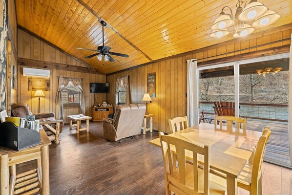 A spacious room with wooden floors, a ceiling fan, a dining table with chairs, a leather sofa, and a large window overlooking a river.