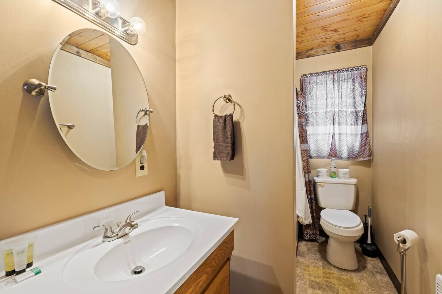 Ein Badezimmer mit rundem Spiegel, Waschbecken, Toilette und einem Fenster mit gemusterten Vorhängen.