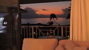 Een balkon met uitzicht op de oceaan bij zonsondergang, een tafel met twee glazen en een fles, en een bank.