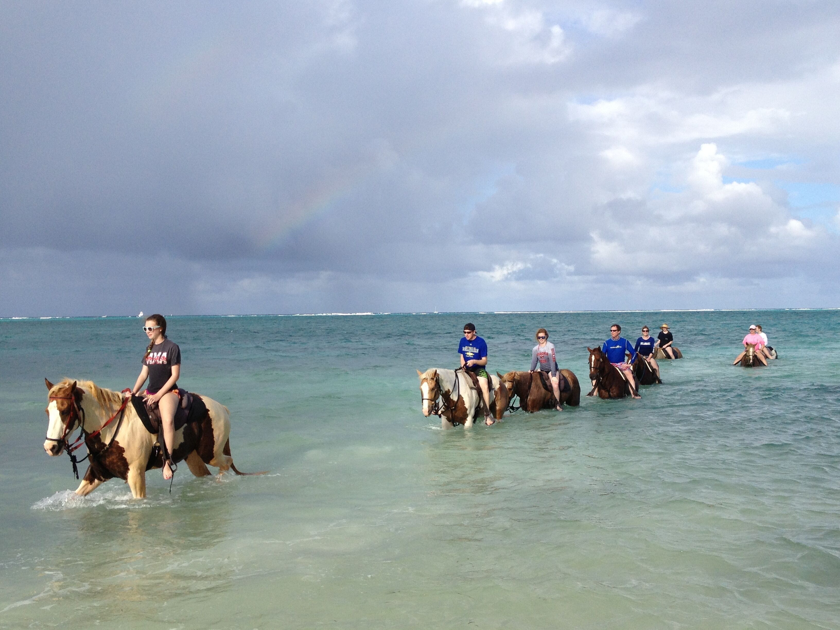 A group of people riding horses in shallow water, with a cloudy sky in the background.