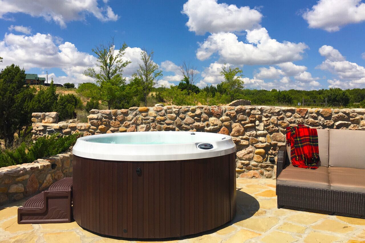A hot tub surrounded by stone walls and greenery, with a wicker sofa and a blanket nearby.