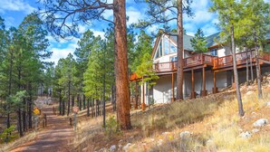 A cabin with a wooden deck and large windows, surrounded by pine trees and a dirt path. - Holland Home - Luxury home w/ Forest Views, AC & Hot Tub! (Flagstaff)
