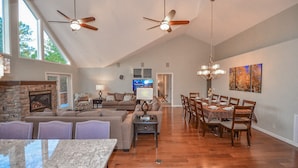A spacious living area with a fireplace, a dining table, and a large window. - Holland Home - Luxury home w/ Forest Views, AC & Hot Tub! (Flagstaff)