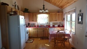 Una cocina con armarios de madera, un refrigerador, una mesa de comedor con sillas y una ventana con cortinas.