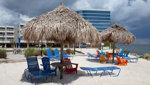 Een strand met rieten parasols, blauwe en rode ligbedden, en een gebouw op de achtergrond.