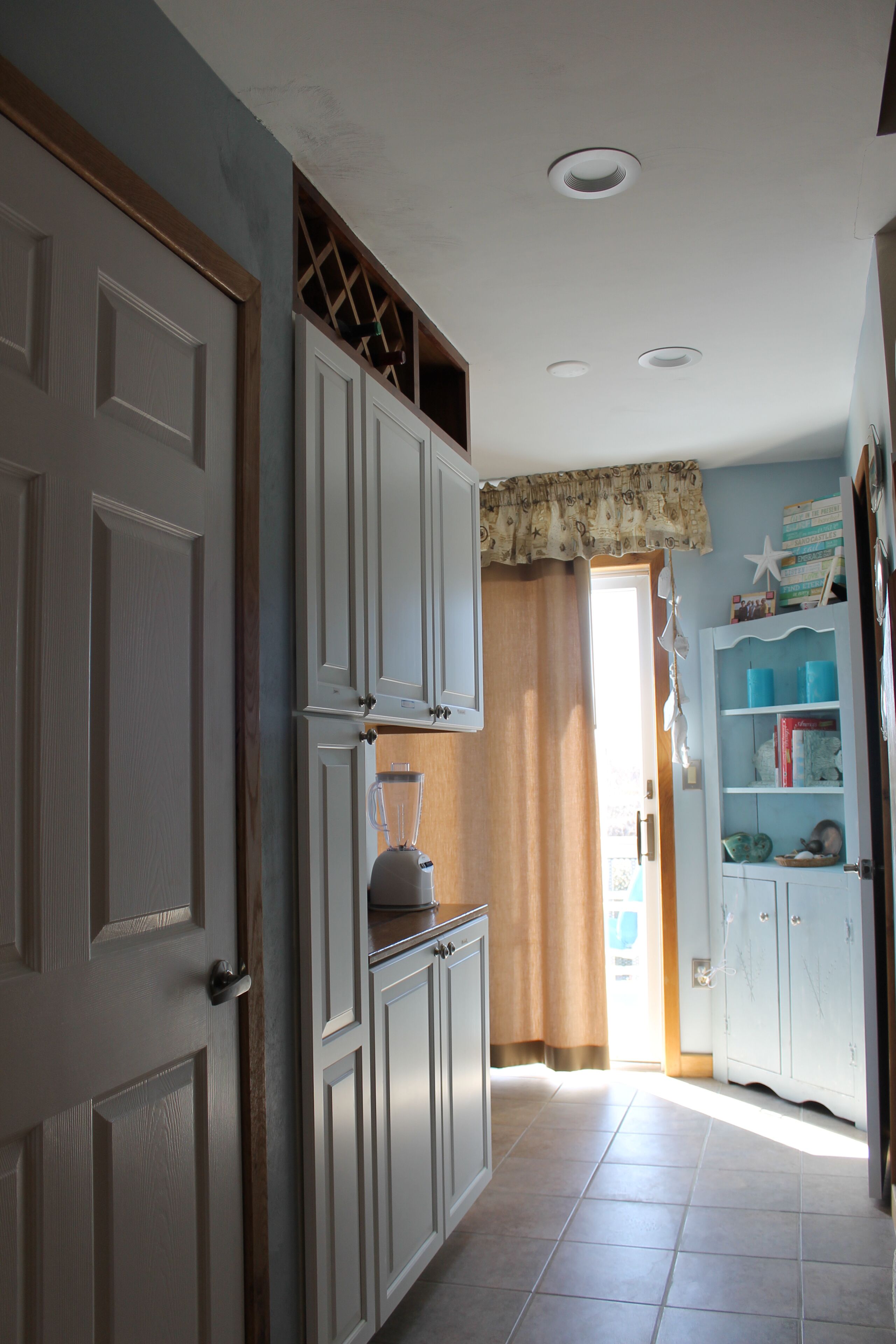A narrow hallway with white cabinets, a door, and a window with curtains.