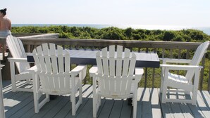 A wooden deck with a table and four white Adirondack chairs, overlooking a body of water and greenery. - Oceanfront property (Duck)