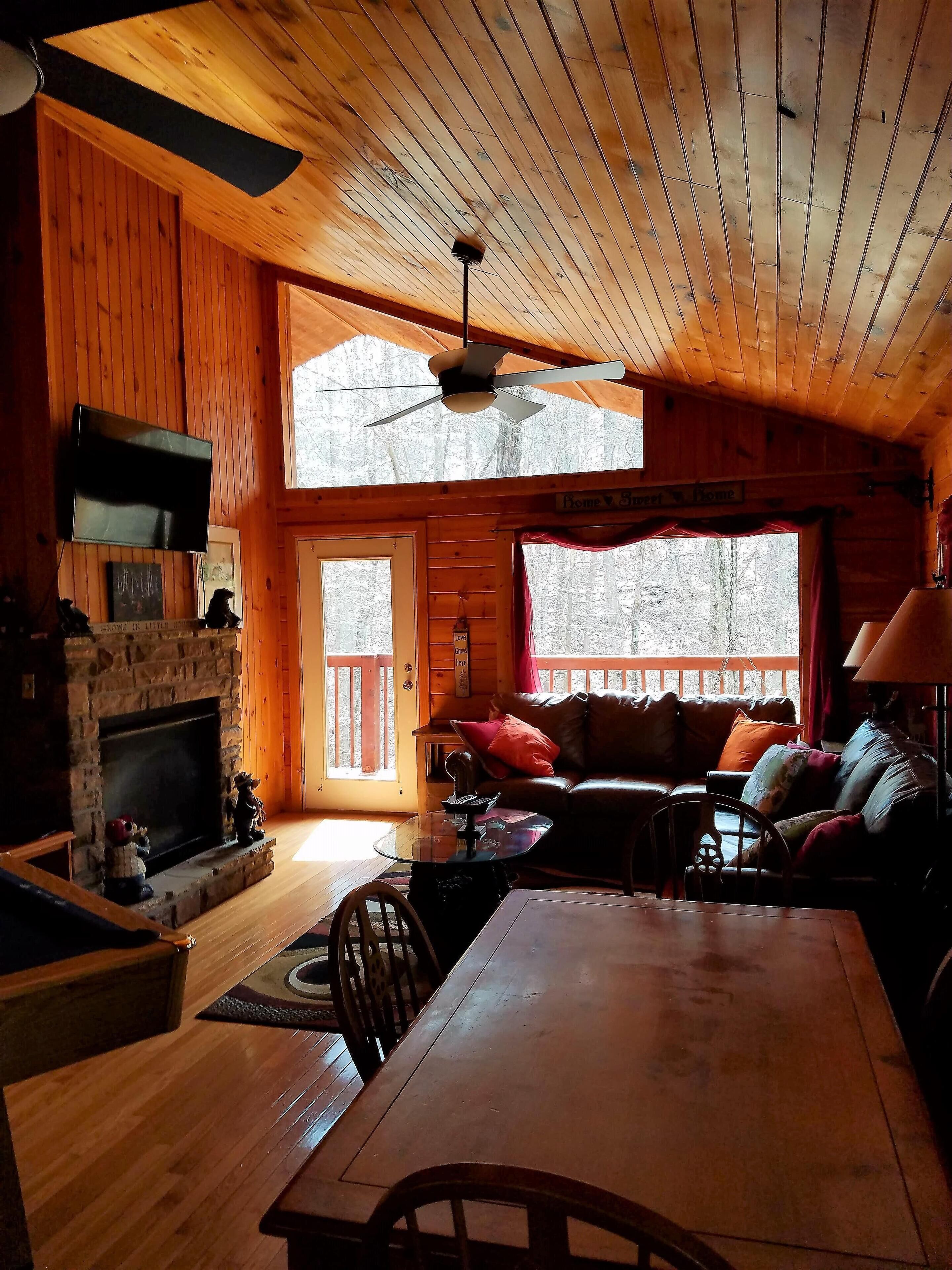 A cozy cabin interior with a stone fireplace, wooden ceiling, a ceiling fan, a television mounted on the wall, and a large window with a view of the outdoors.