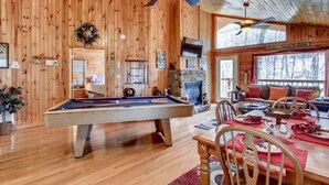 A cozy cabin interior with a pool table, fireplace, and a dining area with a view of the forest.
