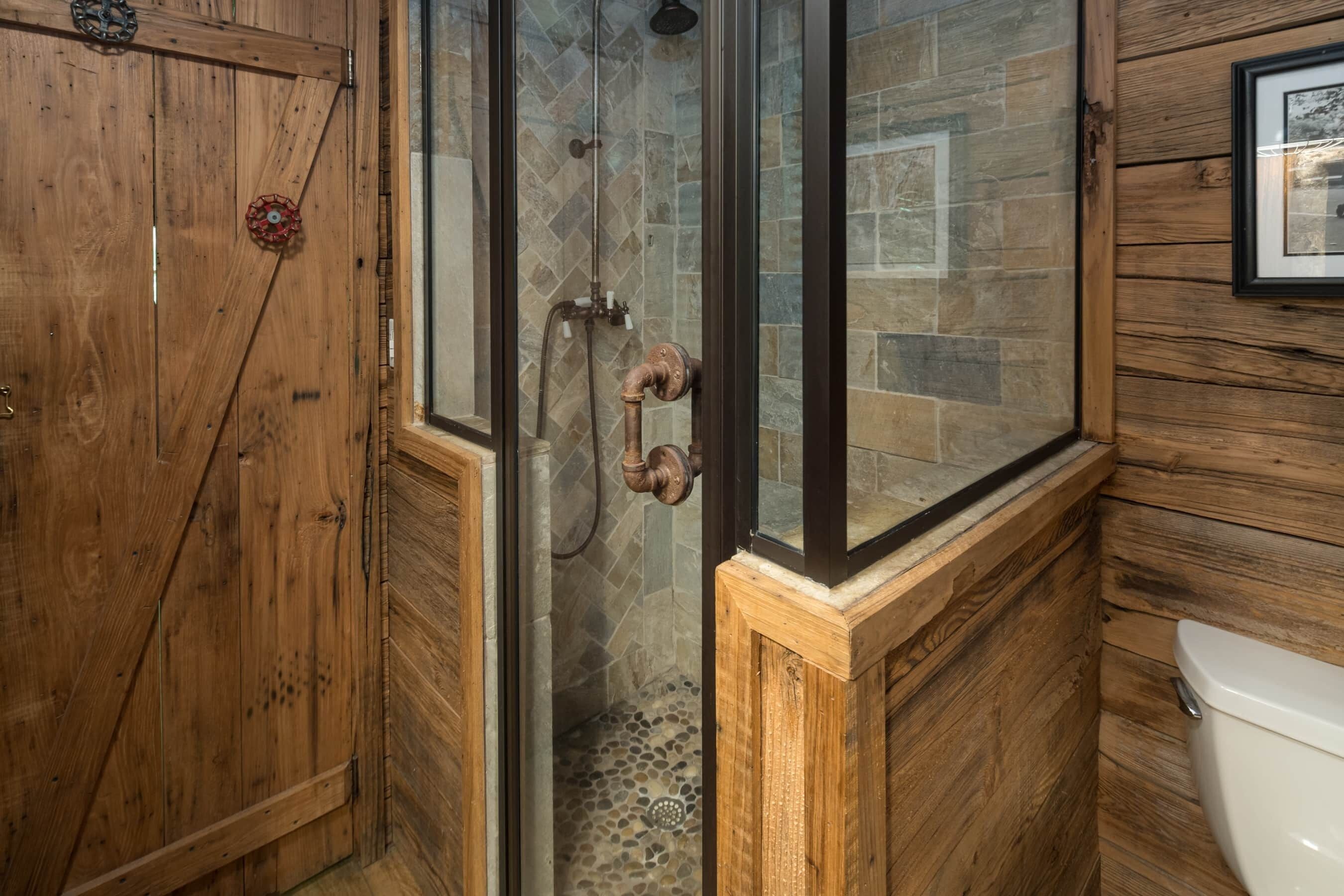 Une salle de bain en bois avec une douche à parois de verre, des toilettes blanches et une porte en bois dotée d’une poignée décorative.