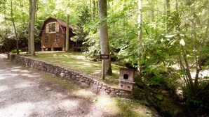 Una cabaña de madera rústica con la señalización "The Barn" rodeada de un bosque denso, con un muro de piedra y un comedero para pájaros cerca.