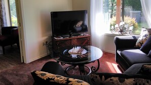 A living room with a television on a stand, a glass coffee table with chess pieces, a sofa, and a window with curtains.
