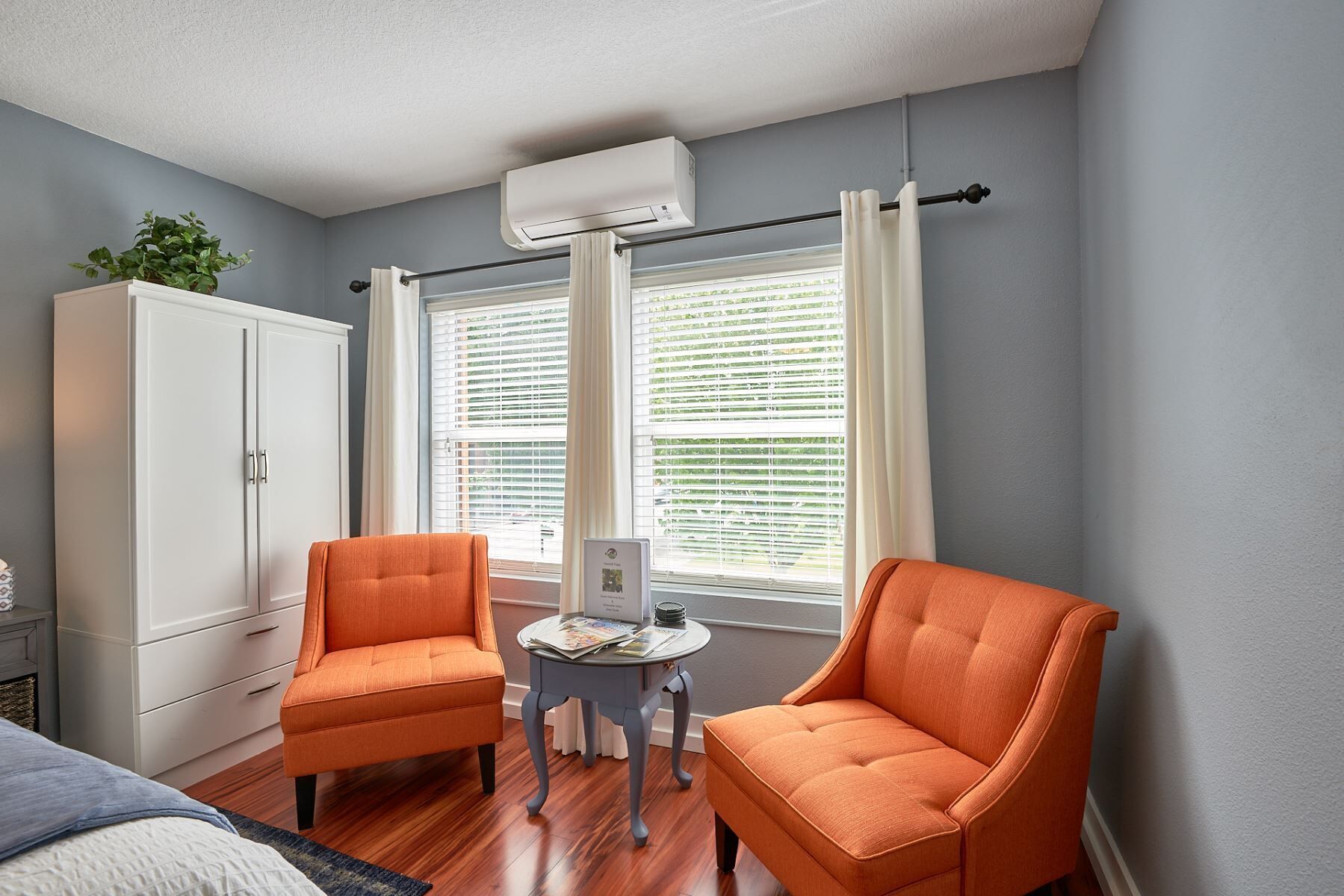 Een kamer met twee oranje fauteuils, een witte kast en een raam met jaloezieën.