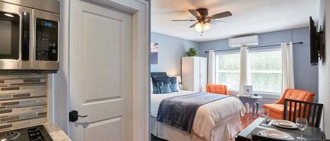 Une chambre d’hôtel moderne dotée d’une kitchenette, d’un lit recouvert d’une courtepointe bleue et blanche, d’un coin repas avec une table dressée pour deux personnes et d’un ventilateur de plafond.