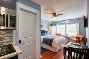 Een moderne hotelkamer met kitchenette, een bed met blauw-witte dekbedovertrek, een eethoek met een tafel gedekt voor twee personen, en een plafondventilator.