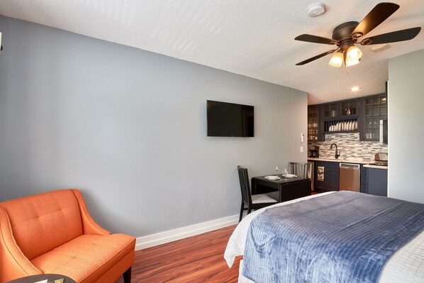 A bedroom with a bed, an orange chair, a desk with a chair, a ceiling fan, and a kitchen area with cabinets and a countertop.