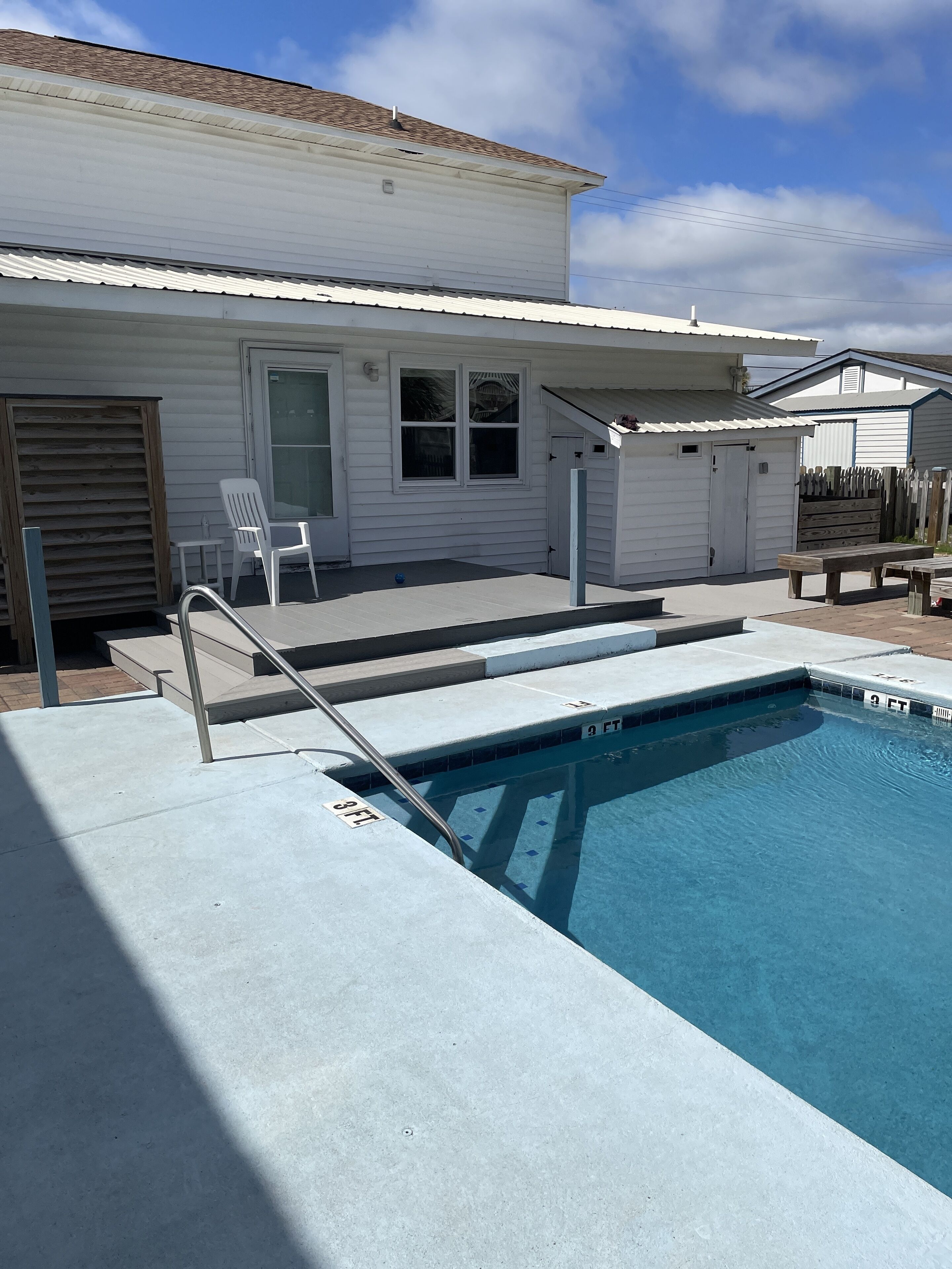 Una piscina residencial con un deck de concreto, una cerca de madera y una casa blanca al fondo.
