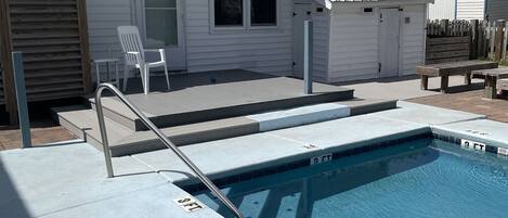 Una piscina residencial con un deck de concreto, una cerca de madera y una casa blanca al fondo.
