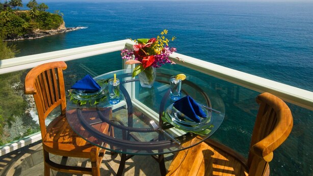 A glass table with two wooden chairs, a vase of flowers, and a glass of water on a balcony with a view of the ocean. - Stunning, majestic, oceanfront location, with stunning view and air conditioning (Hilo)