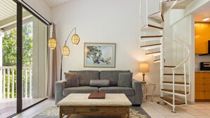 Una sala de estar con un sofá gris, una mesa de centro, una silla de mimbre, un cuadro en la pared y una escalera en espiral.