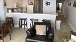 A living room with a black leather sofa, wooden chairs, and a small table. A kitchen with white cabinets and a refrigerator is visible in the background. - JUST LISTED, OPEN CALENDAR, FRIENDLY PRICES (Panama City Beach)