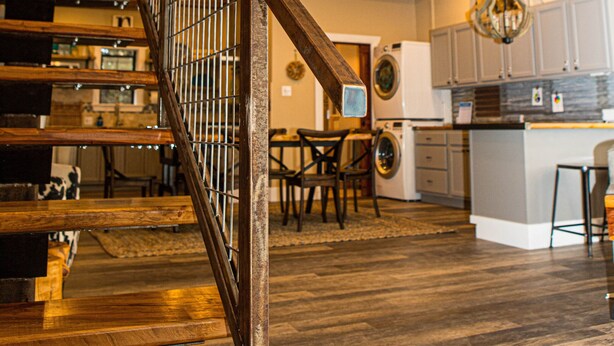 Una escalera de madera de varios niveles con barandillas metálicas, una zona de cocina con armarios y electrodomésticos, y un comedor con sillas y mesas.