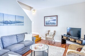 A living room with a grey sofa, a wicker chair, a glass coffee table, a TV on a stand, and a staircase.