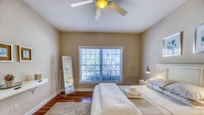 Une chambre à coucher comprenant un lit, une table de chevet, une commode, un ventilateur de plafond, une fenêtre et une étagère murale ornée de cadres avec des photos.