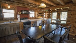 A rustic cabin-style kitchen with wooden beams, a central island, and a dining table with benches. - Authentic Log cabin with something for everyone! (Marthasville)