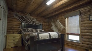 A wooden cabin bedroom with a bed, a dresser, a zebra-patterned pillow, a large animal hide, and a window with blinds. - Authentic Log cabin with something for everyone! (Marthasville)
