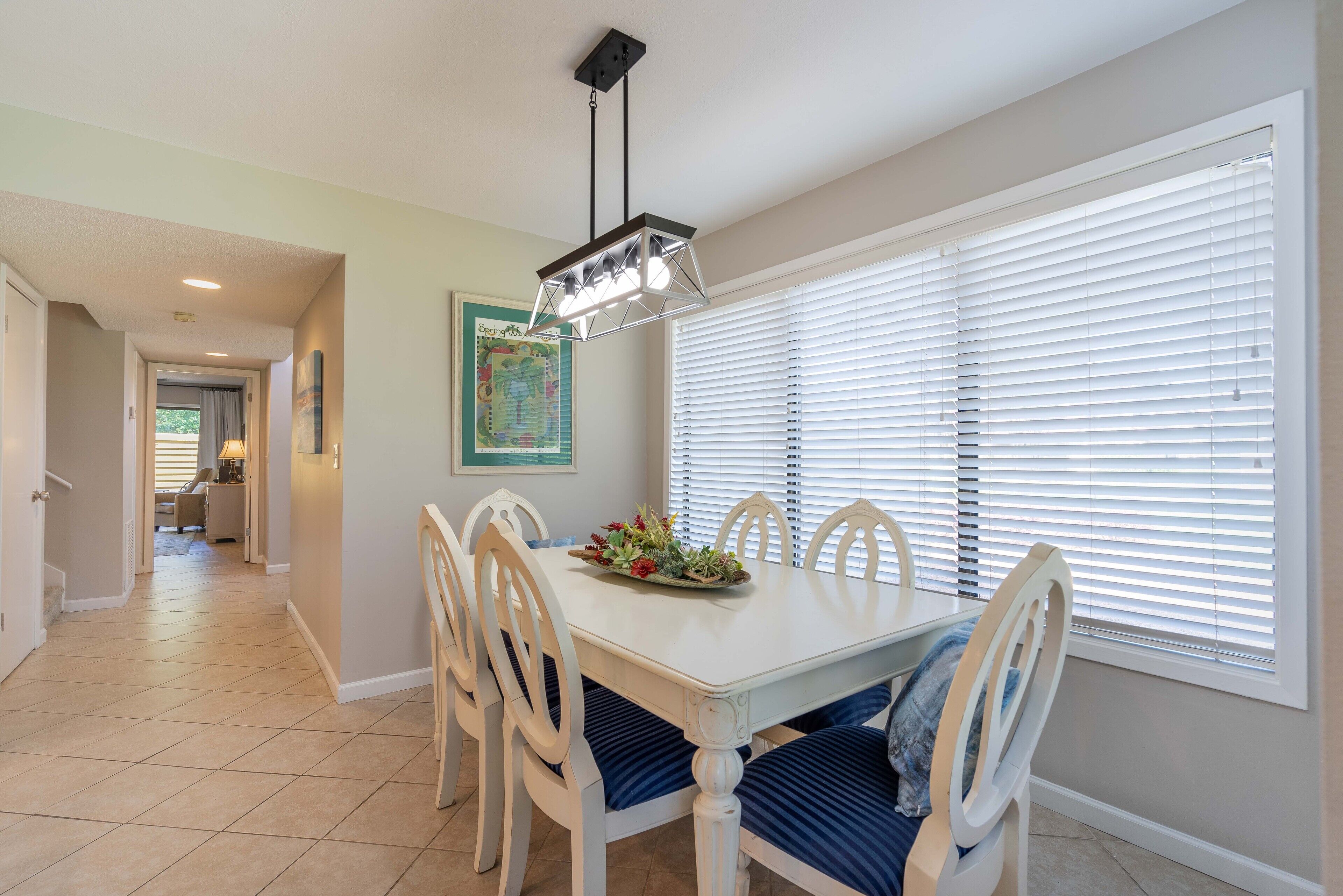 A dining area with a white table, blue cushioned chairs, a hanging light fixture, and a window with blinds.
