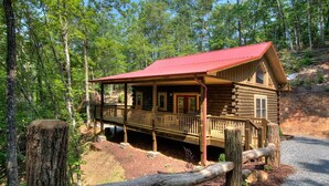 A log cabin with a red roof, wooden deck, and a porch.