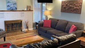 Een woonkamer met een bank, een salontafel, een open haard, een televisie en kunstwerken aan de wanden.
