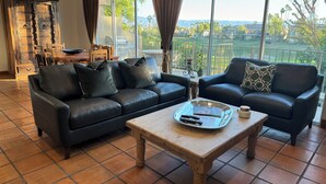 A living room with a black leather sofa, a wooden coffee table, and a view of a green landscape through large windows. - PGA West La Quinta STR #068246 - Close to Golf, Tennis & Music Festivals (La Quinta)
