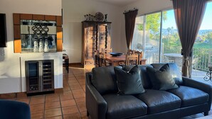 A living room with a black leather sofa, a dining table with chairs, a wooden cabinet, and a large window with curtains. - PGA West La Quinta STR #068246 - Close to Golf, Tennis & Music Festivals (La Quinta)