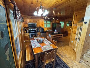A cabin interior with a wooden dining table, chairs, a kitchen with appliances, and a ceiling light fixture.