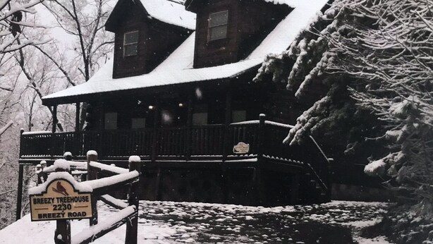 A wooden house with a snow-covered roof and a sign reading "Breezy Treehouse 2230 Breezy Road."