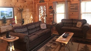 A cozy cabin-style room with wooden walls and a large TV on the left. There are two sofas with bear-themed cushions and a small table with a lamp.