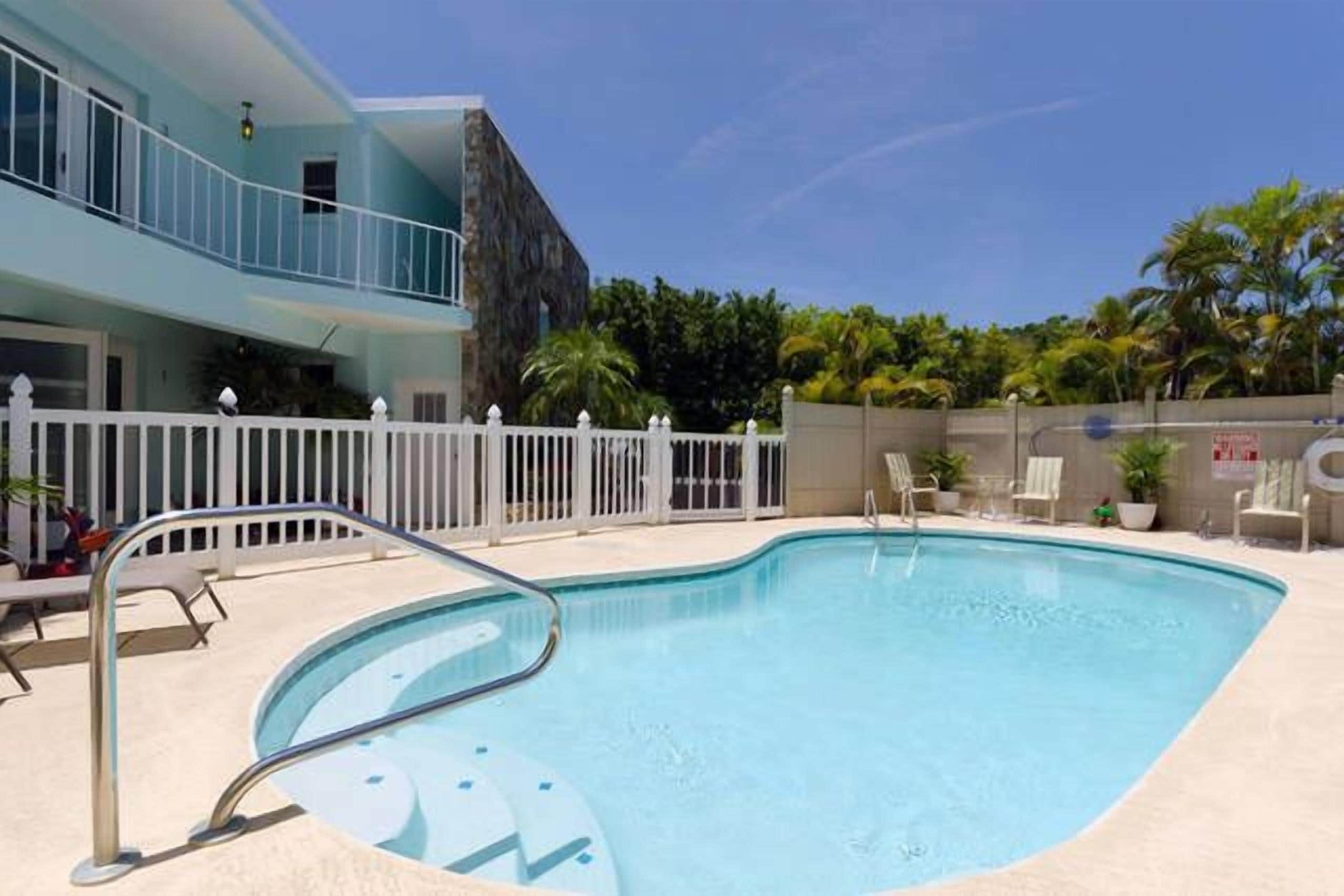 A swimming pool with a white fence, lounge chairs, and a building with a balcony.