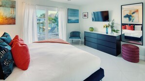 Un dormitorio moderno con una cama grande, un escritorio, una silla, un televisor y vistas a una piscina y edificios.