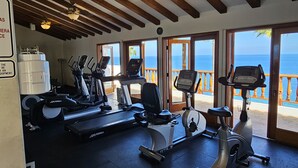 Een sportschool met diverse fitnessapparaten, een groot waterreservoir en uitzicht op de oceaan door grote deuren.