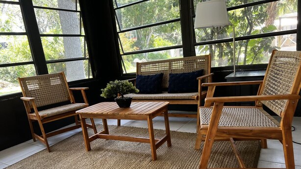 Una habitación con un ventanal grande, mobiliario de mimbre, una mesita con una planta y una lámpara.