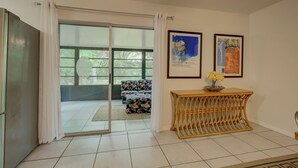 Una habitación con puerta de vidrio corredera que da a un balcón, una mesita de madera con un jarrón de flores y dos cuadros en la pared.