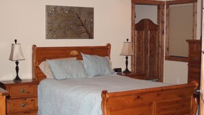 Ein hölzernes Bett mit Kopfteil, zwei Nachttische mit Lampen, ein Nachttisch mit Schubladen, ein Holzschrank und ein an der Wand befestigtes Kunstwerk.